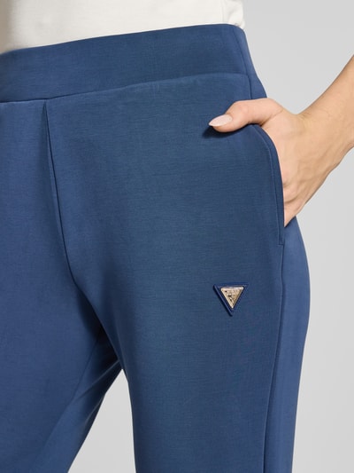 Kobieta nosi Sweatpants mit Label-Detail od Guess Activewear, niebieski elastyczny materiał, proste nogawki, boczne kieszenie, detal z logo, zbliżenie na biodro.