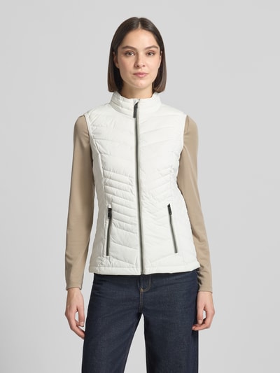 Vrouwenmodel draagt bodywarmer met 10.000 mm waterkolom model 'GS109' van G.I.G.A. DX, slim fit, opstaande kraag, ritssluiting, ritszakken, ruglengte bij maat 36: 61 cm, schouderbreedte: 37 cm, borstwijdte: 49 cm, licht beige kleur, vooraanzicht volledige lichaam.