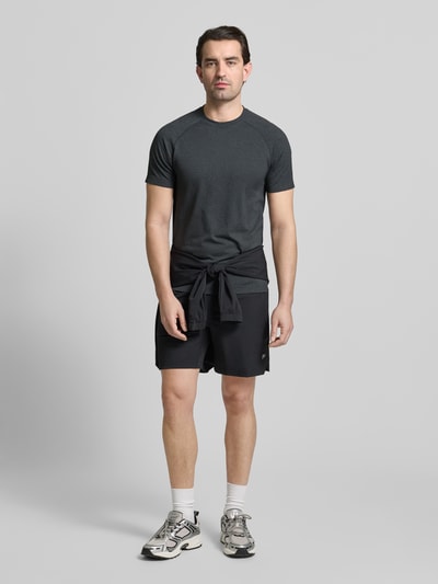 Męski model nosi Regular Fit T-Shirt mit Rundhalsausschnitt od Fabletics, logo, krótki rękaw, ciemny kolor, krój regular fit, widok z przodu, ujęcie całej sylwetki
