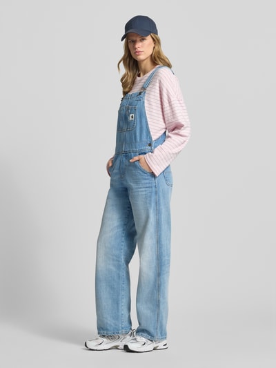 Vrouwenmodel draagt Latzhose met borstzak en labelpatch van Carhartt Work In Progress, puur katoen, straight fit, verstelbare bandjes met haakjessluiting, borstzak, labelpatch, zijsteekzakken, achterzakken, ruglengte maat S 32 cm, borstwijdte maat S 40 cm, totale lengte maat S 133 cm, binnenbeenlengte maat S 78 cm, lichtblauw denim, casual uitstraling, voorkant geheel in beeld