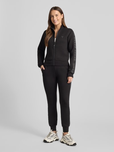 Frauenmodel trägt Regular Fit Sweatjacke aus Viskose-Mix von Guess Activewear in Schwarz mit Logo-Applikation und elastischen Details, Ganzkörperansicht von vorne.