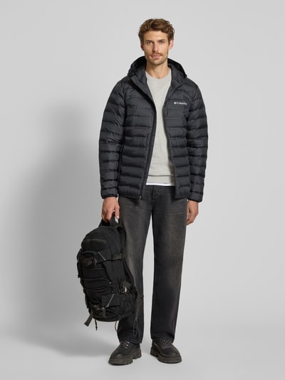 Męskamodel nosi czarną kurtkę pikowaną z efektem pikowania, krój regular fit, kaptur, nadruk z logo, zamek błyskawiczny, Model 'Lake 22™' od Columbia, kurtka przejściowa, Długość tyłu w rozmiarze M: 72 cm, Szerokość ramion w rozmiarze M: 43 cm, Szerokość klatki piersiowej w rozmiarze M: 42 cm, Długość rękawów w rozmiarze M: 70 cm, zdjęcie całej sylwetki z przodu.