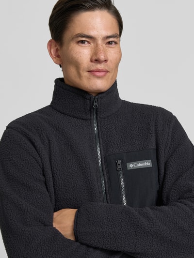 Männermodel trägt Fleecejacke mit Stehkragen Modell 'Jasper' von Columbia in Anthrazit mit Brusttasche und Label-Patch, Nahaufnahme Oberkörper. Gerader Schnitt, durchgehender Reißverschluss, seitliche Eingrifftaschen, elastische Ärmelabschlüsse.