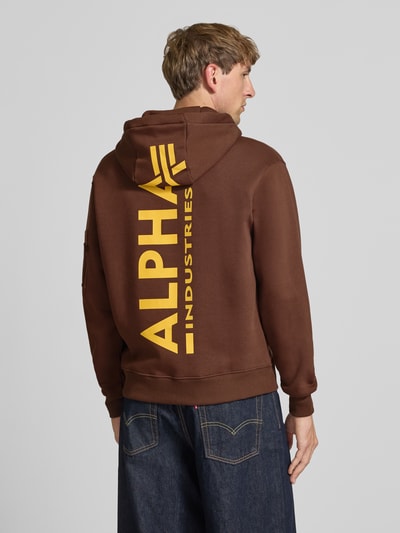 Männermodel trägt Hoodie mit Känguru-Tasche von Alpha Industries in Braun mit gelbem Label-Print, Baumwoll-Mix, lockerer Schnitt, Ganzkörperansicht Rückansicht.