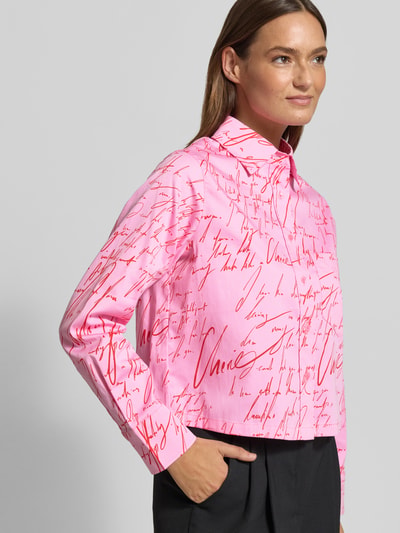 Modelka nosi Regular Fit Hemdbluse aus Baumwoll-Mix von s.Oliver BLACK LABEL, efekt: wzór na całej powierzchni, mieszanka bawełny, koszulowy kołnierzyk, długi rękaw, mankiet, kieszeń na piersi, listwa guzikowa, pas na plecach z zakładką czeladniczą, kolor różowy z czerwonym napisem, całość z przodu do pasa, z przodu.