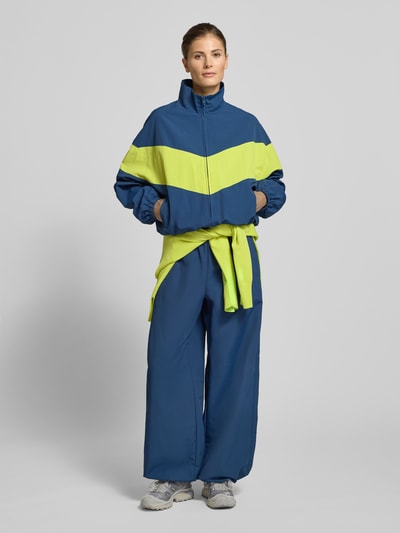 Vrouwenmodel draagt Nylon-Jacke im 90s-Stil - curated by Tinvcb van Tinvcb in blauw met felgele strepen, regular fit, hoge kraag, ritssluiting, elastische boorden, steekzakken met ritssluiting; ruglengte 60 cm, schouderbreedte 54 cm, borstwijdte 58 cm, mouwlengte 34 cm; frontaal volledige lichaamsopname.