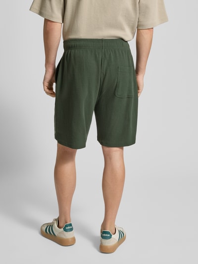Männermodel trägt Shorts mit Logo-Print und Eingrifftaschen von ANOTHER COTTON LAB, dunkelgrün, lockerer Schnitt, Rückansicht Ganzkörperansicht.