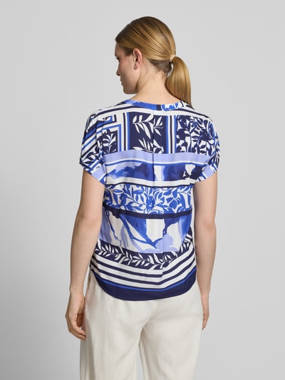 Frauenmodel trägt Blusenshirt aus Viskose mit Strasssteinbesatz von Betty Barclay, Motiv- und Statement-Print in Blau- und Weißtönen, Straight Fit, Tunikakragen, Kappärmel, Rückenlänge 55 cm, Ganzkörperansicht Rückansicht.