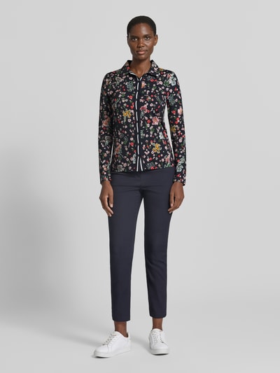 Frauenmodel trägt Slim Fit Hemdbluse mit floralem Allover-Print von Marc Cain, dunkle Grundfarbe, bunte Blumenmuster, elastischer Material-Mix, Hemdblusenkragen, langärmlig, glatte Textur, kombinierbar, Ganzkörperansicht von vorne.