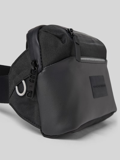 Produktfoto, Tasche mit Label-Detail von Porsche Design in Schwarz mit reflektierenden Streifen, wasserdicht, Hauptfach mit Reißverschluss, längenverstellbarer Schulterriemen, Maße: 23 cm Breite, 14 cm Länge, 8 cm Tiefe.