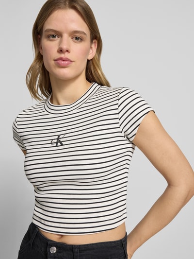 Modelka nosi Cropped T-Shirt mit Streifenmuster und Logo von Calvin Klein Jeans, mieszanka bawełny i elastanu, wzór w paski, logo, okrągły dekolt, damski t-shirt, krój krótki, długość tyłu w rozmiarze S: 39 cm, kolor czarno-biały, faktura prążkowana, widok na górną część ciała z przodu