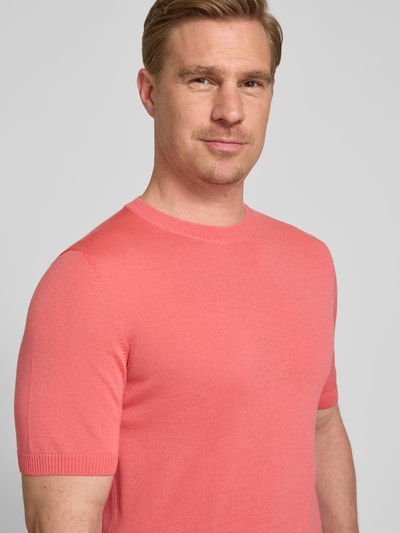 Mannelijk model draagt Strickshirt mit Rundhalsausschnitt van MAERZ Muenchen, slim fit, ribboorden, labeldetail, korrelige textuur, zalmroze kleur, detailopname bovenlichaam.