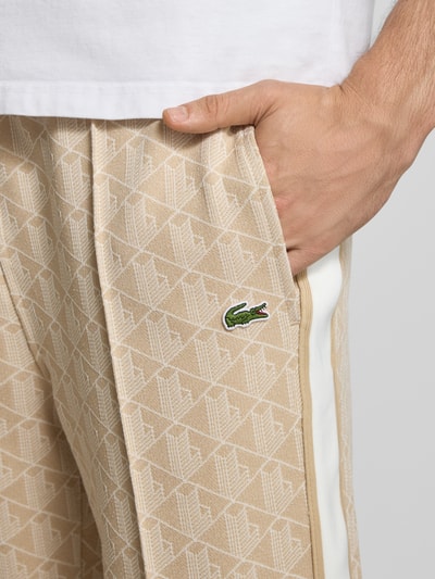 Mężczyzna nosi Regular Fit Sweathose mit Baumwoll-Anteil od Lacoste, wzór na całej powierzchni, naszywka z logo, paski w kontrastowym kolorze, wpuszczane kieszenie, dzianina dresowa, beżowa i biała, detailaufnahme.