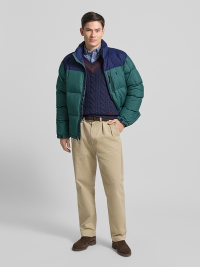 Męski model nosi Kurtka puchowa z zamkiem błyskawicznym dwustronnym i stójką od Polo Ralph Lauren, wypełnienie puchem i pierzem, krój regular fit, wyhaftowane logo, stójka, kieszeń na piersi, długość tyłu 73 cm, szerokość ramion 50 cm, szerokość klatki piersiowej 57 cm, długość rękawów 70 cm, całość w odcieniach zieleni i granatu, tekstura pikowana, gładkie spodnie, brązowe buty, całkowity widok z przodu.