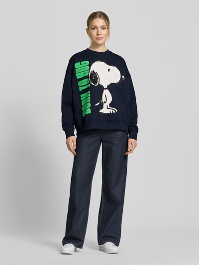 Frauenmodel trägt Loose Fit Sweatshirt aus reiner Baumwolle Modell 'JUMBRELLA EMBO' von Essentiel Antwerp in Dunkelblau mit Snoopy-Motiv, Pailletten und Perlen, lange Ärmel, gerippte Abschlüsse, Ganzkörperansicht von vorne.