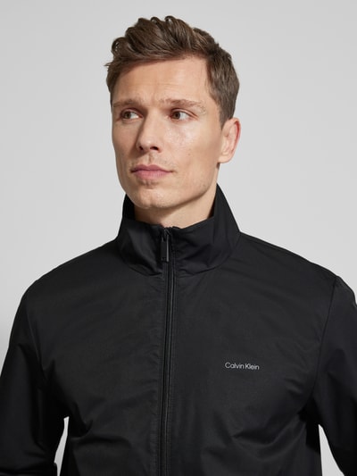 Männermodel trägt Jacke mit Stehkragen und Reißverschluss von CK Calvin Klein in Schwarz, glatte Textur, Nahaufnahme Oberkörper, Regular Fit.