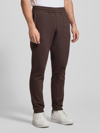 Männermodel trägt Sweatpants mit elastischem Bund van adidas Originals in bruin, katoenmix, los model, steekzakken, labelstitching, ganzkörperansicht van voren.