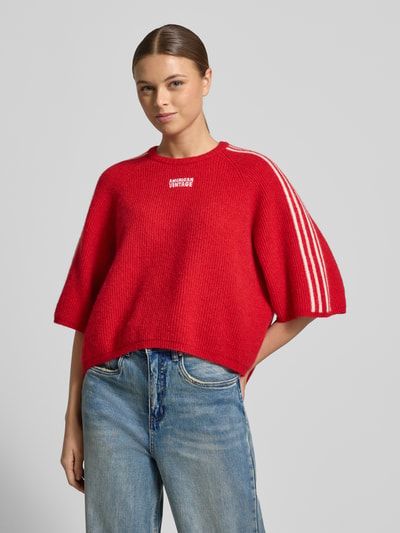 Frauenmodel trägt Cropped Strickpullover mit gerippten Abschlüssen Modell 'VITOW' von American Vintage, rot mit weißen Details, Ganzkörperansicht von vorne. Damen-Strickpullover aus Woll-Alpaka-Mix mit Stretch, lockerer Schnitt, Rundhalsausschnitt, Label-Stitching, Rückenlänge 53 cm, Ärmellänge 36 cm.
