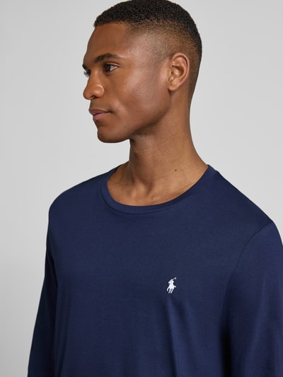 Męski model nosi bluzkę z długim rękawem z bawełny z wyhaftowanym logo od Polo Ralph Lauren, prosty krój, okrągły dekolt, zbliżenie na górę sylwetki.