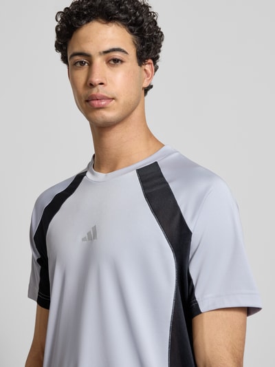 Męski model nosi T-Shirt mit Label-Print von adidas Training w kolorze szarym z czarnymi wstawkami, krój regular fit, ściągacz przy okrągłym dekolcie, długość tyłu w rozmiarze M: 72 cm, zbliżenie na górną część ciała z przodu.