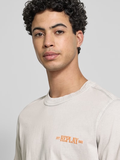 Mężczyzna nosi Regular Fit T-Shirt mit Logo-Print od Replay z bawełny, kolor jasny beż, okrągły dekolt, nadruk z przodu, zbliżenie na górę ciała
