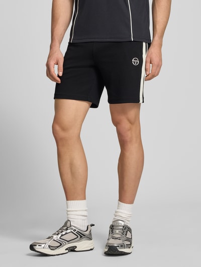 Męski model nosi czarne szorty z wyhaftowanym logo, mieszanka bawełny z dodatkiem streczu, wpuszczane kieszenie, luźny krój, model 'EGEO' od SERGIO TACCHINI, elastyczny pas z trokiem; szerokość w rozmiarze M: 40 cm, długość wewnętrzna nogawki w rozmiarze M: 20 cm; widok całej sylwetki od przodu