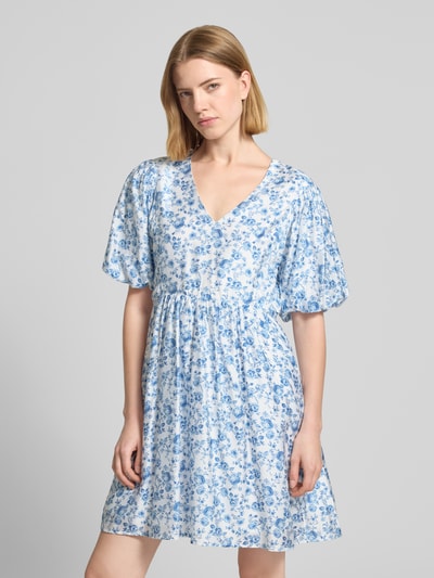 Frauenmodel trägt tailliertes Freizeitkleid aus Viskose-Mix Modell 'BLISS' von Vero Moda, Optik:Allover-Muster, Mini-Länge, V-Ausschnitt, kurze Ballonärmel, Saum gerade, Materialverarbeitung: feine Webstruktur, Farben: Weiß mit blauem Muster, Ganzkörperansicht von vorne.