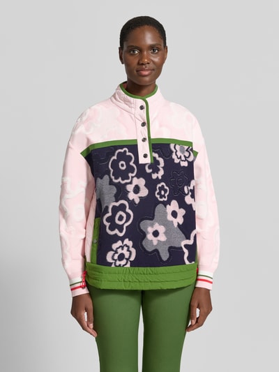 Frauenmodel trägt Relaxed Fit Pullover aus Viskose-Mix mit Troyer-Kragen von Marc Cain, florales Design in Rosa, Dunkelblau und Grün, Rückenlänge 62 cm, Ärmellänge 54 cm, Ganzkörperansicht von vorne.