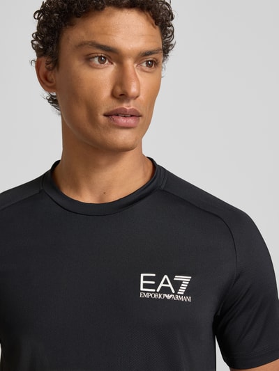 Männermodel trägt T-Shirt mit Label-Print von EA7 Emporio Armani in Schwarz, Material-Mix mit Stretch-Anteil, Nahaufnahme Oberkörper.