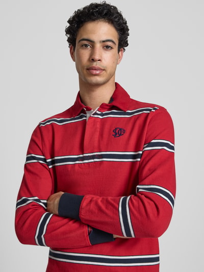 Männermodel trägt Regular Fit Poloshirt mit Logo-Stitching von Superdry in Rot mit navy-weißen Streifen, Reine Baumwolle, Nahaufnahme Oberkörper. Rückenlänge bei Größe M: 71 cm, Polokragen, Knopfleiste, Label-Details.