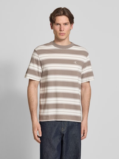 Männermodel trägt Regular Fit T-shirt aus katoenmix van Calvin Klein Jeans met gestreept all-over motief en gestikt logo, ronde hals, korte mouw, geheel in neutrale tinten, ganzkörperansicht van voren.