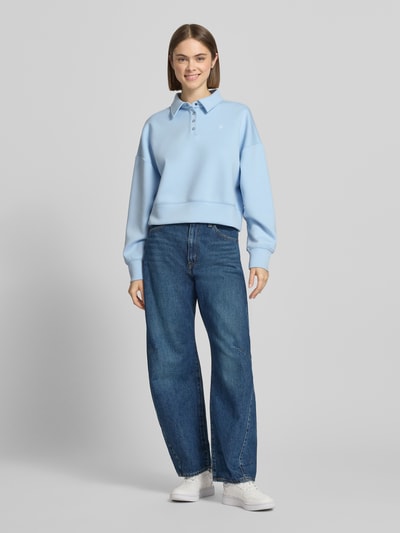 Frauenmodel trägt Cropped Pullover mit Viskose-Anteil von Guess Jeans in Hellblau, Polo-Kragen, Logo-Stitching, Ganzkörperansicht von vorne.