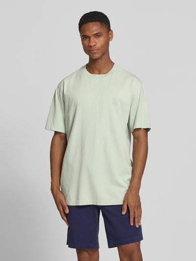 Männermodel trägt Relaxed Fit T-Shirt aus reiner Bio-Baumwolle Modell 'ICONIC MAARKOS GD' von Armedangels in hellem Grün, unifarben, kurzarm, Jersey-Textur, Logo-Print, Ganzkörperansicht von vorne.