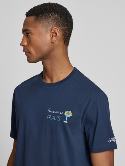 Męski model nosi t-shirt z haftami model 'PORTOFINO' od MC2 Saint Barth, bawełna, krój regular fit, okrągły dekolt, długość tyłu w rozmiarze M: 68 cm, kolor granatowy, tekstura gładka. Zbliżenie na górną część ciała z boku.