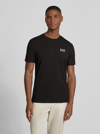 Männermodel trägt schwarzes T-Shirt mit Label-Print von EA7 Emporio Armani, Slim Fit, Baumwoll-Elasthan, Ganzkörperansicht von vorne.