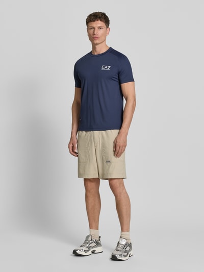 Männermodel trägt T-Shirt mit Label-Print von EA7 Emporio Armani in Dunkelblau, Slim Fit, Material-Mix, Ganzkörperansicht von vorne.
