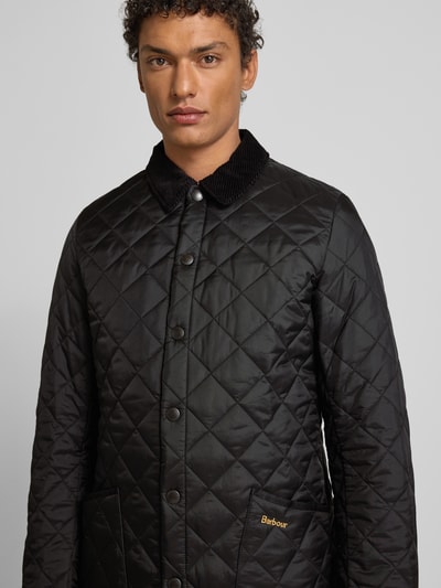 Męski model nosi Steppjacke mit Umlegekragen von Barbour, krój regular fit, jednokolorowy, pikowanie, wykładany kołnierz, listwa z zatrzaskami, wpuszczane kieszenie, wyhaftowane logo, czarny kolor, długość tyłu 68 cm, szerokość ramion 40 cm, szerokość klatki piersiowej 42 cm, długość rękawów 70 cm, półzbliżenie z przodu.