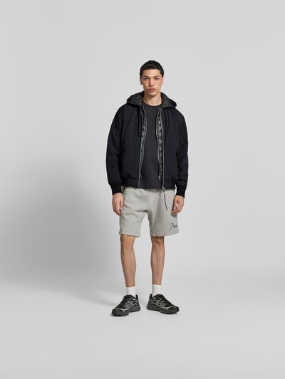 Männermodel trägt Bomberjacke mit Daunen-Feder-Füllung von Moose Knuckles in Schwarz, lockere Passform, Kapuze mit Tunnelzug, Zweiwege-Reißverschluss, Känguru-Tasche mit Patten, kombiniert mit grauer Shorts und schwarzen Sneakern, Farben neutral, glatte und matte Textur, Ganzkörperansicht von vorne. Rückenlänge 63 cm, Schulterbreite 48 cm, Ärmellänge 65 cm.
