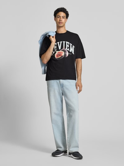 Männermodel trägt Relaxed Fit T-Shirt mit Logo-Print von REVIEW in Schwarz, Reine Baumwolle, Rundhalsausschnitt, Halbarm, Rückenlänge 68 cm, Ganzkörperansicht von vorne.