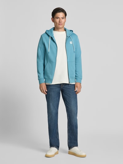 Männermodel trägt Sweatjacke mit Logo-Patch und Känguru-Tasche von Scotch & Soda in Blau, Frontansicht, Kontext: reine Baumwolle, gerader Schnitt, Kapuze mit Kordelzug, Reißverschluss, gerippte Abschlüsse, Rückenlänge 70 cm, Ärmellänge 68 cm, Ganzkörperansicht von vorne.