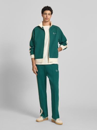 Männermodel trägt Regular Fit Sweatjacke mit Stehkragen von KARL KANI in Grün mit Logo-Stitching, Reißverschluss, Elastischer-Material-Mix, Eingrifftaschen, elastische Abschlüsse, Rückenlänge und Ärmellänge bei Größe M, Ganzkörperansicht von vorne.