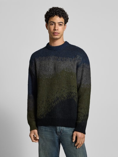 Männermodel trägt Strickpullover mit Rundhalsausschnitt Modell 'TINLEY' von Carhartt Work In Progress in Blau und Oliv, Material-Mix, Ganzkörperansicht von vorne.