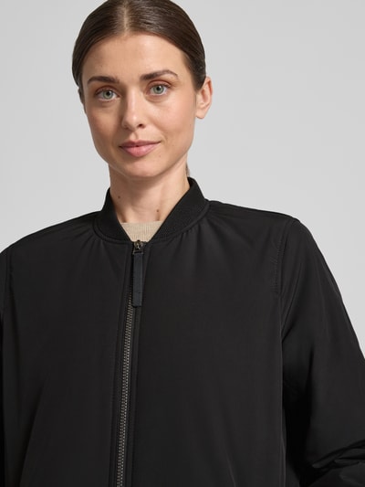 Kobiece modelka nosi Kurtka 'Jacke mit Zweiwege-Reißverschluss' od camel active, czarna, stójka, luźny krój, zamek błyskawiczny dwustronny, elastyczne zakończenia rękawów, bardzo długi krój, Nahaufnahme górnej części ciała.