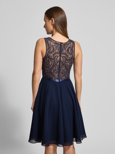 Kobiece modelka nosi sukienkę koktajlową z obszyciem z kamieni stras Cocktailkleid mit Strasssteinbesatz od LUXUAR FASHION, półprzezroczysty materiał, lekko taliowany fason, długość do kolan, jednokolorowy design, zamek błyskawiczny, okrągły dekolt, długość tyłu 96 cm, szerokość klatki piersiowej 41 cm, widok całej sylwetki z tyłu.