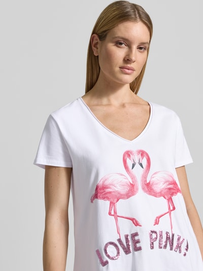 Modelka kobieca nosi damski t-shirt z mieszanki bawełny i elastanu, krój regular fit, nadruk z motywem, model 'Flamingo Love Me' od miss goodlife, dekolt w serek, długość tyłu w rozmiarze S: 66 cm, motyw flaminga w odcieniach różu, napis love pink, tekstura gładka, półzbliżenie na górną część ciała.