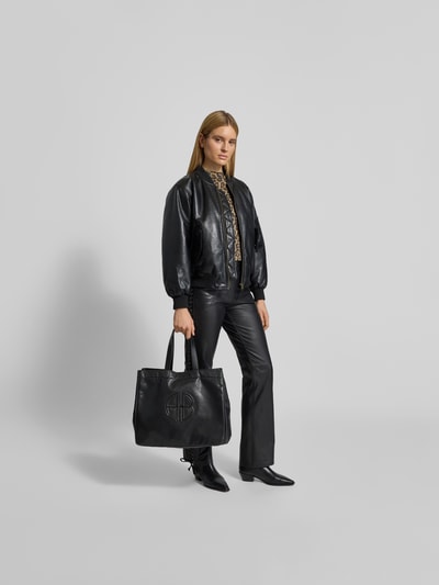 Frauenmodel trägt schwarze Tote Bag mit Logo-Detail von Anine Bing, Strukturmuster, Magnetverschluss, Schulterriemen, Ganzkörperansicht von vorne.