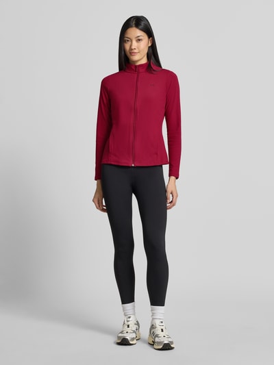 Frauenmodel trägt Slim Fit Trainingsjacke mit Logo-Print von Under Armour in Rot, kombiniert mit schwarzer Leggings, Ganzkörperansicht von vorne, Damen Trainingsjacke von Under Armour.