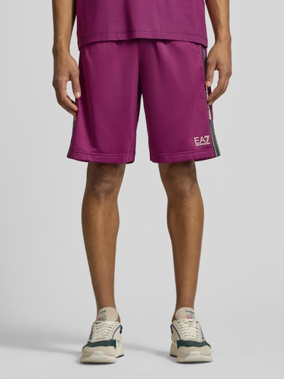 Męski model nosi Shorts mit Logo und Eingrifftaschen von EA7 Emporio Armani, fuksjowe, krój relaxed fit, szerokość w rozmiarze M: 42 cm, długość wewnętrzna nogawki: 28 cm, wpuszczane kieszenie, logo, elastyczny pas, całkowity widok z przodu.