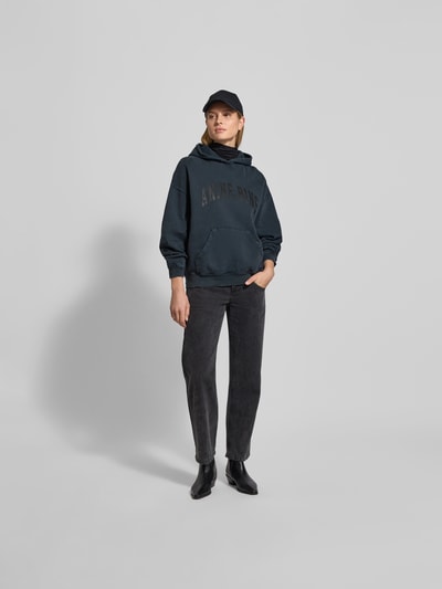 Frauenmodel trägt Oversized Hoodie mit Logo-Stitching von Anine Bing aus reiner Baumwolle, Oversized-Fit, Känguru-Tasche, elastische Abschlüsse, Rückenlänge bei Größe S: 63 cm, dunkle Farben, weiche Textur, Ganzkörperansicht von vorne.