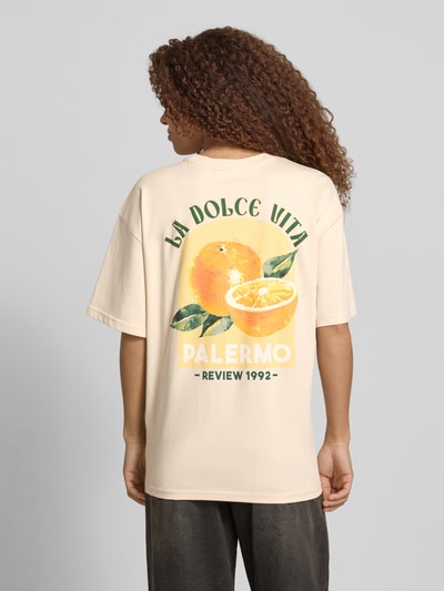 Frauenmodel trägt T-Shirt mit rückseitigem Motiv-Print von REVIEW in Beige mit Rundhalsausschnitt, Oversized, reine Baumwolle, Rückenlänge 68 cm, Ganzkörperansicht Rückansicht.